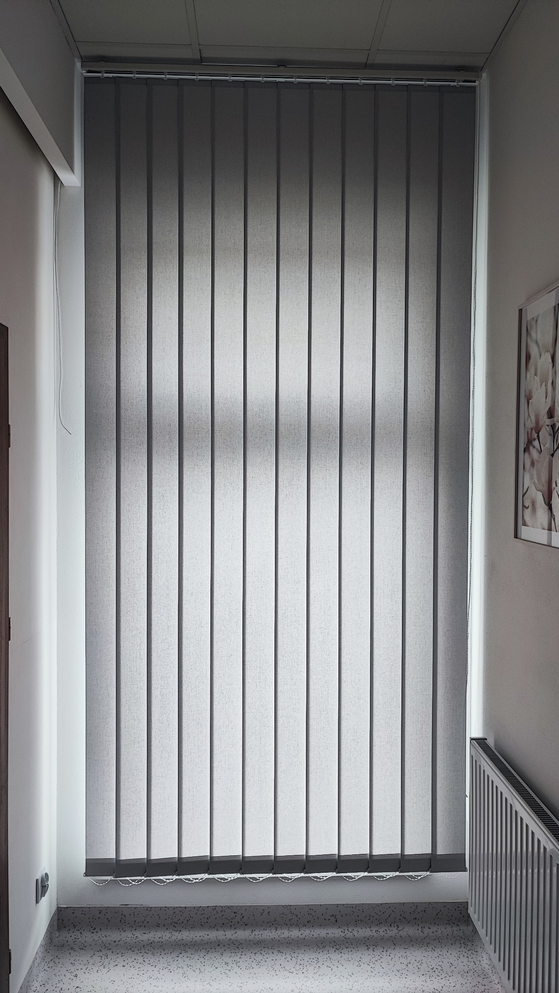 Szare żaluzje pionowe (wertykale) zamontowane w pomieszczeniu, widoczny fragment grzejnika i obrazka na ścianie.