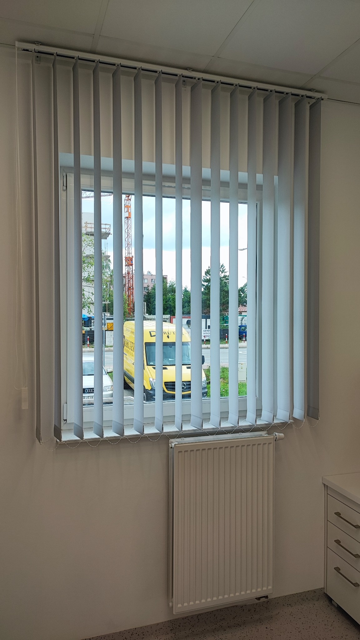 Wnętrze biura z widokiem na ulicę przez okno z szarymi żaluzjami pionowymi; za oknem widoczny żółty samochód dostawczy z logo firmy na tle budowy z dźwigiem.