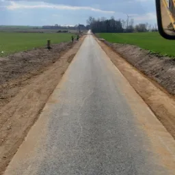 Świeżo utwardzona droga gruntowa widziana z kabiny maszyny budowlanej, z dwoma pracownikami w oddali, zielone pola po bokach i wiatrak na horyzoncie pod pochmurnym niebem.
