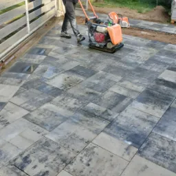 Pracownik zagęszcza wibroprasą nawierzchnię z szaro-czarnych kostek brukowych na posesji z białym płotem i przyczepą kempingową w tle.