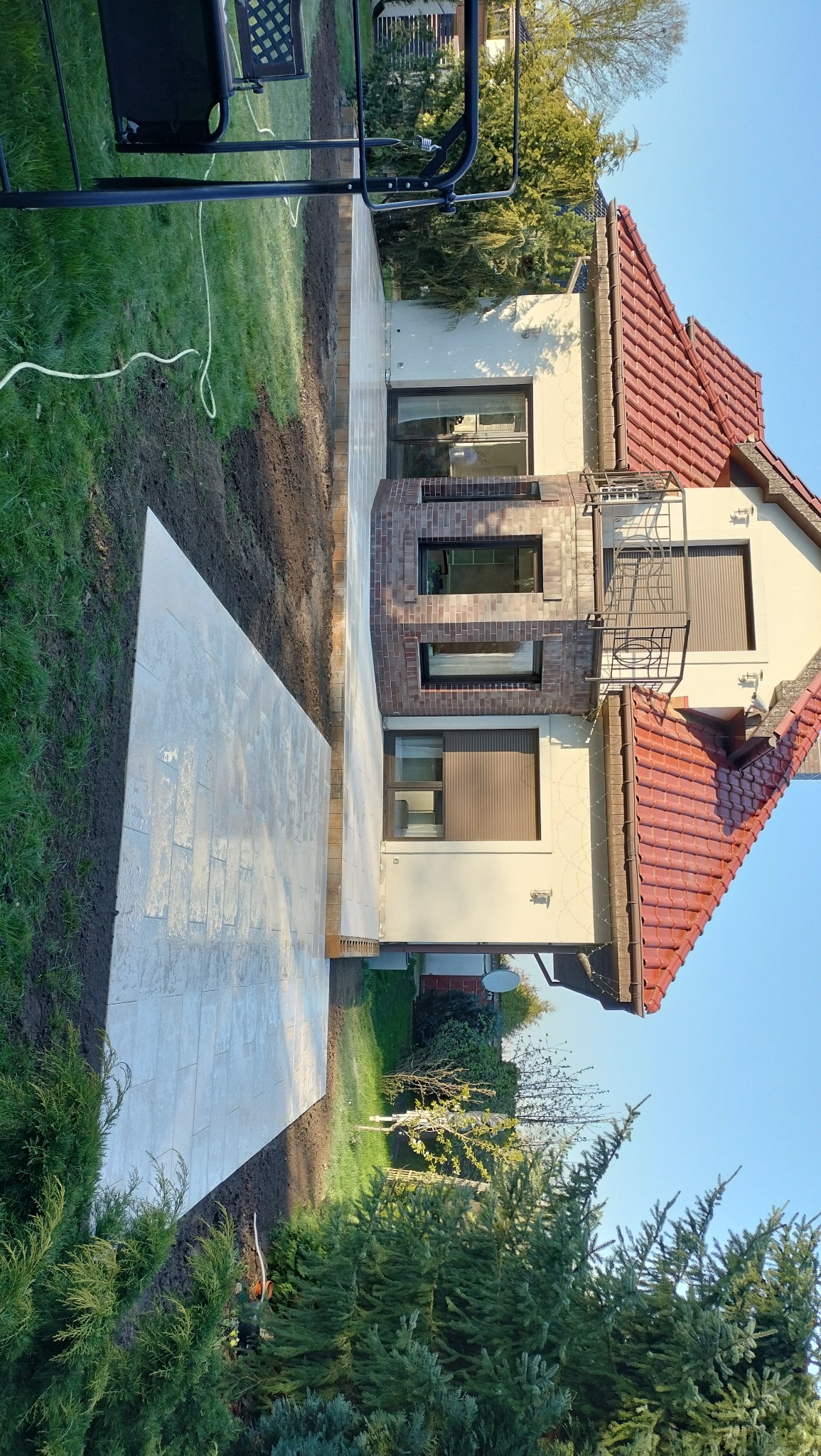 Ułożona jasna nawierzchnia z płyt w ogrodzie przylegająca do domu z czerwoną dachówką, świeżo obsypana ziemią, widoczny trawnik i roślinność.