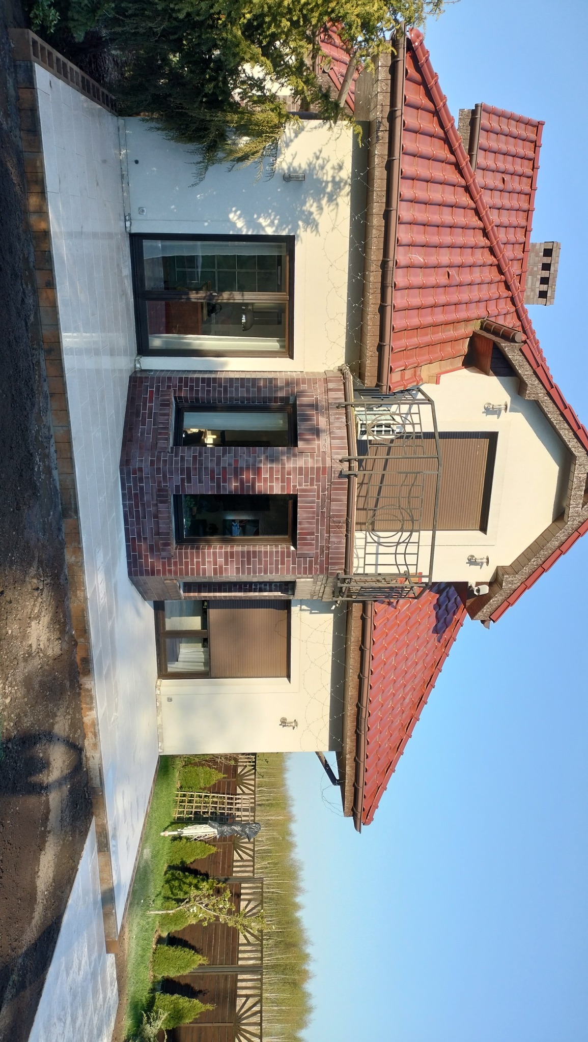 Wykończony fragment elewacji domu z nową, jasną nawierzchnią z kostki brukowej, ceglanym wykuszem i balkonem z metalową balustradą, w tle widoczny trawnik i ogrodzenie.