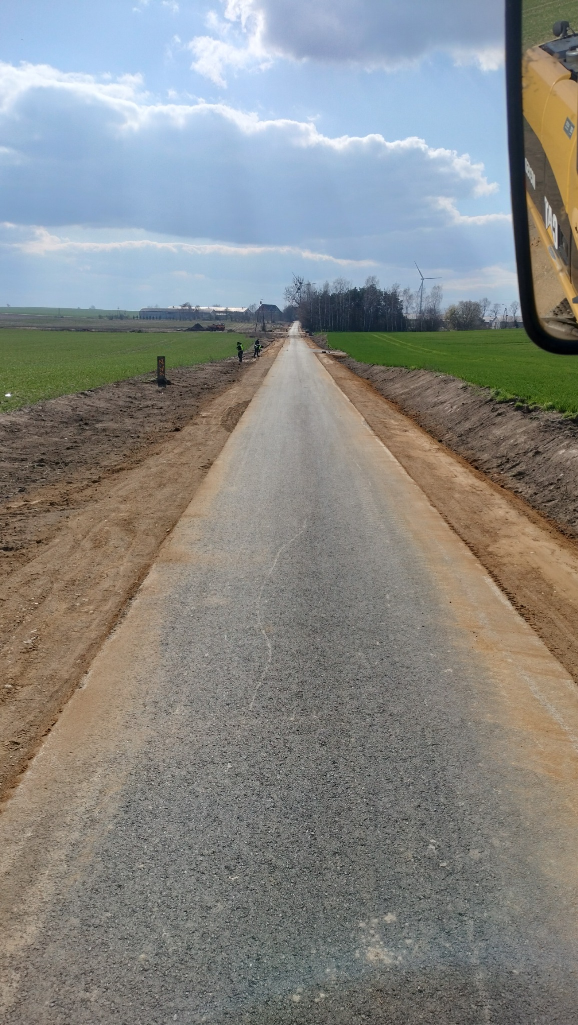 Świeżo utwardzona droga gruntowa widziana z kabiny maszyny budowlanej, z dwoma pracownikami w oddali, zielone pola po bokach i wiatrak na horyzoncie pod pochmurnym niebem.