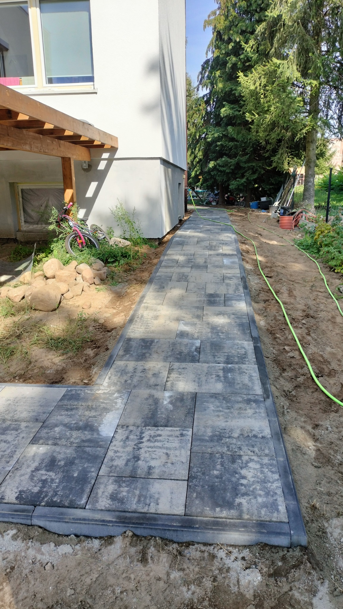 Ścieżka z ułożonej, szarej kostki brukowej z ciemną obwódką, prowadząca wzdłuż budynku z widocznymi elementami ogrodu i zieleni w tle.