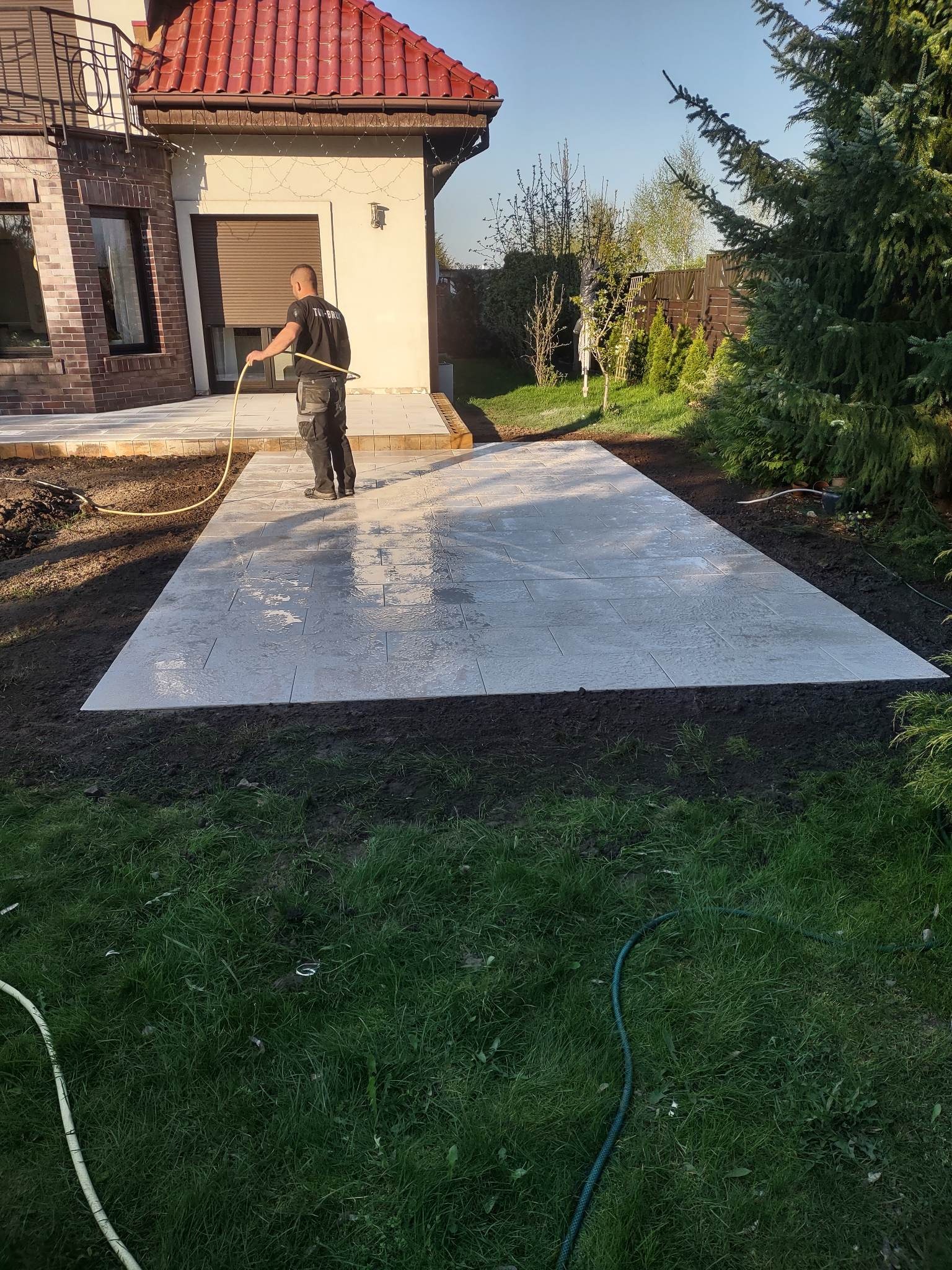 Pracownik spryskuje wodą świeżo ułożony taras z szarej kostki brukowej na posesji z domem w tle.