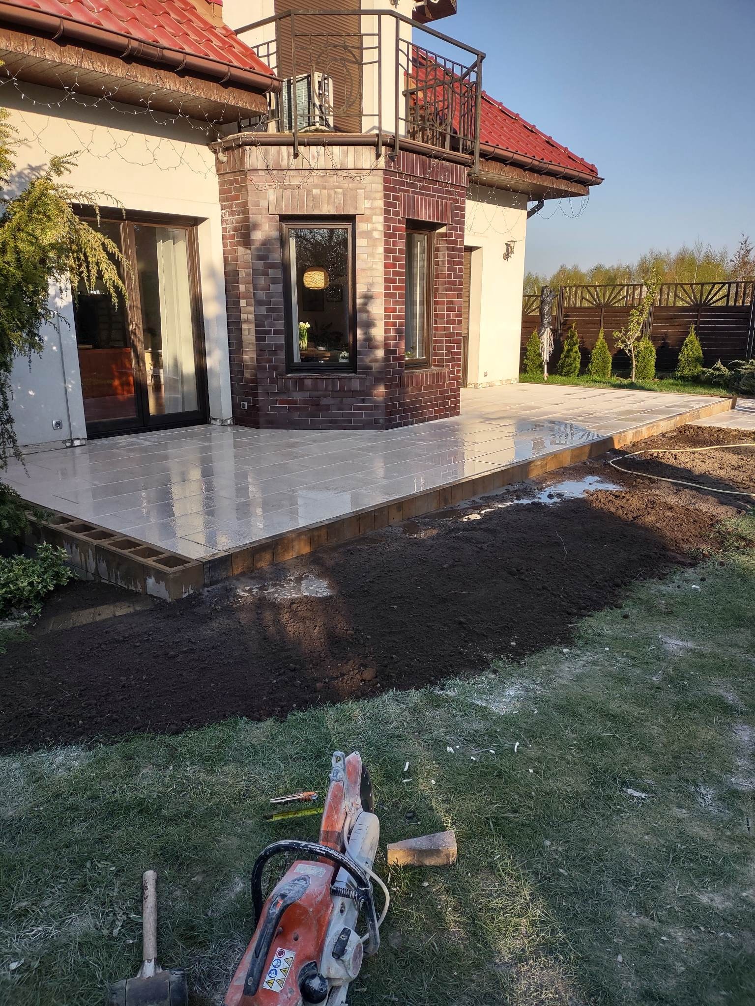 Jasne płyty tarasowe ułożone przy domu z ceglanym elementem, widoczne narzędzia brukarskie na trawie, przygotowany teren pod dalsze prace.