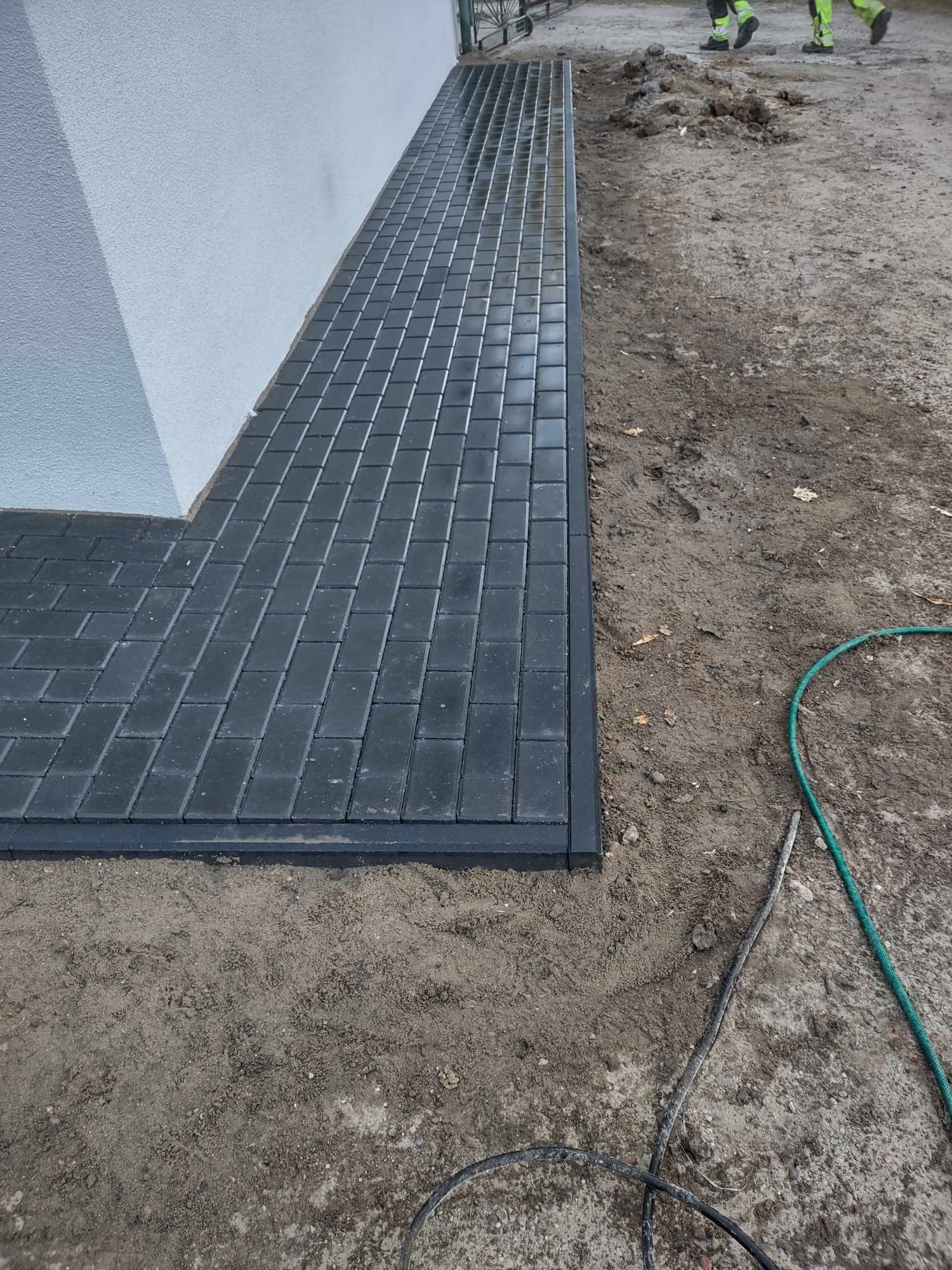 Ułożona, ciemnoszara kostka brukowa tworząca ścieżkę przy ścianie budynku. Widoczne obrzeża kostki, ziemia po bokach i fragment węża ogrodowego. W tle pracownicy w odblaskowych ubraniach.