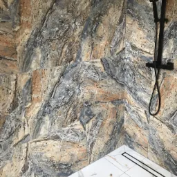 Wykończona łazienka z marmurowymi płytkami ściennymi w odcieniach beżu, szarości i brązu, białą marmurową podłogą z czarnym odpływem liniowym i czarną armaturą prysznicową.