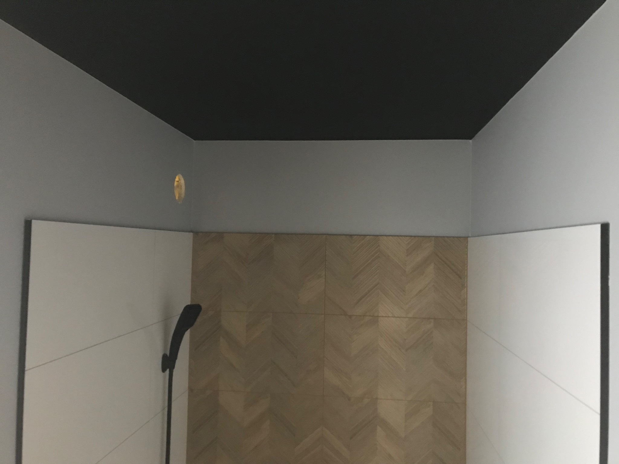 Nowoczesna łazienka z czarnym sufitem, szarymi ścianami i płytkami w jodełkę na ścianie prysznica, widoczna czarna słuchawka prysznicowa.