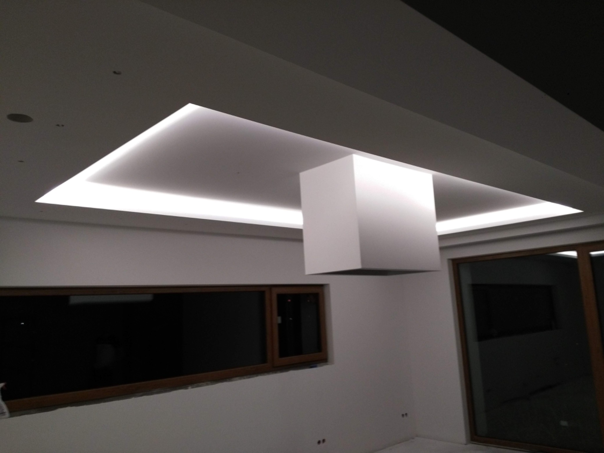 Nowoczesne wnętrze z podwieszonym sufitem z oświetleniem LED w kształcie kwadratu i centralną kolumną, widoczne okna z ciemnymi szybami i surowy stan wykończenia podłogi.