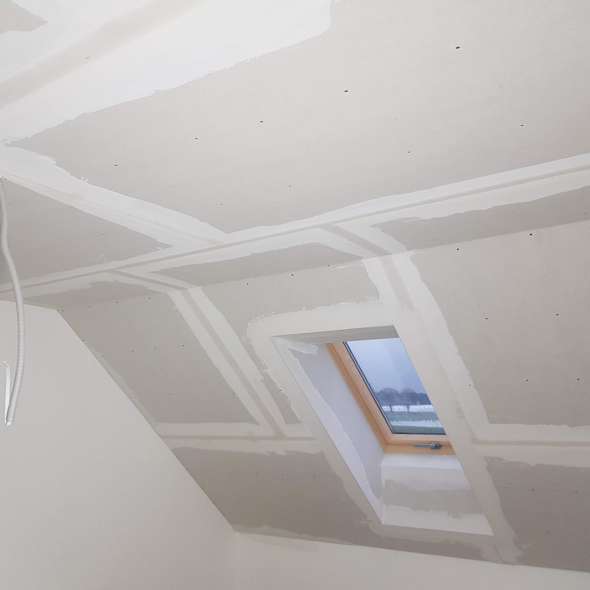 Poddasze z oknem dachowym w trakcie szpachlowania płyt gipsowo-kartonowych, widoczne łączenia płyt i otwory po wkrętach, zarys zimowego krajobrazu za oknem.