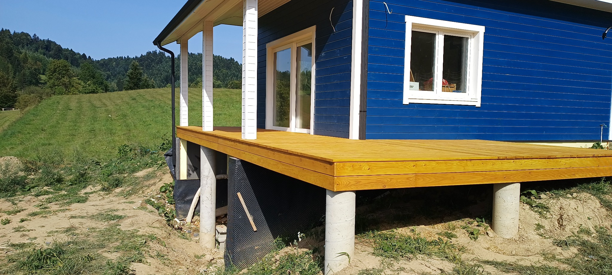 Niebieski dom z drewnianym tarasem na betonowych podporach, widok z boku na tle zielonego wzgórza.