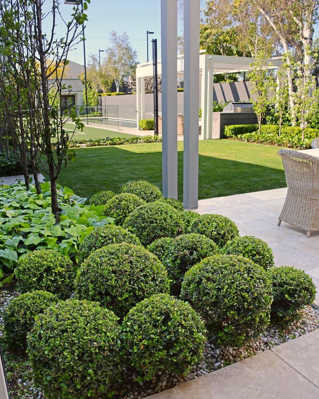 Starannie utrzymany ogród z kulistymi krzewami na żwirowym podłożu, równo przystrzyżony trawnik w tle i nowoczesna pergola.
