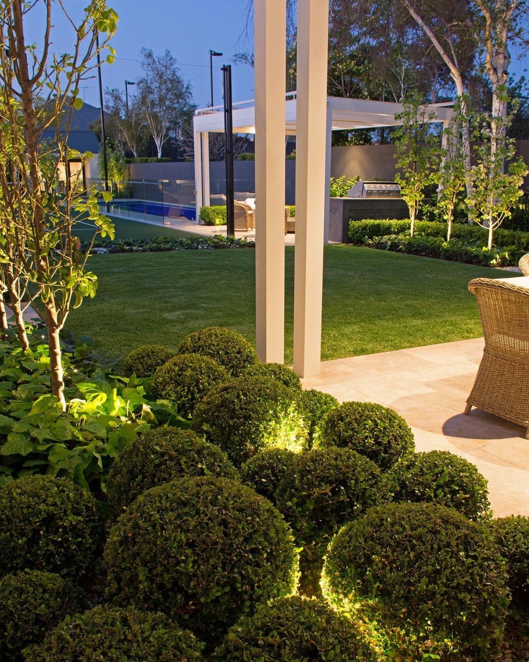 Ogród z oświetleniem punktowym podkreślającym kuliste krzewy, równo przystrzyżony trawnik, basen z szklanym ogrodzeniem i biała pergola w tle, oświetlona wieczorną porą.