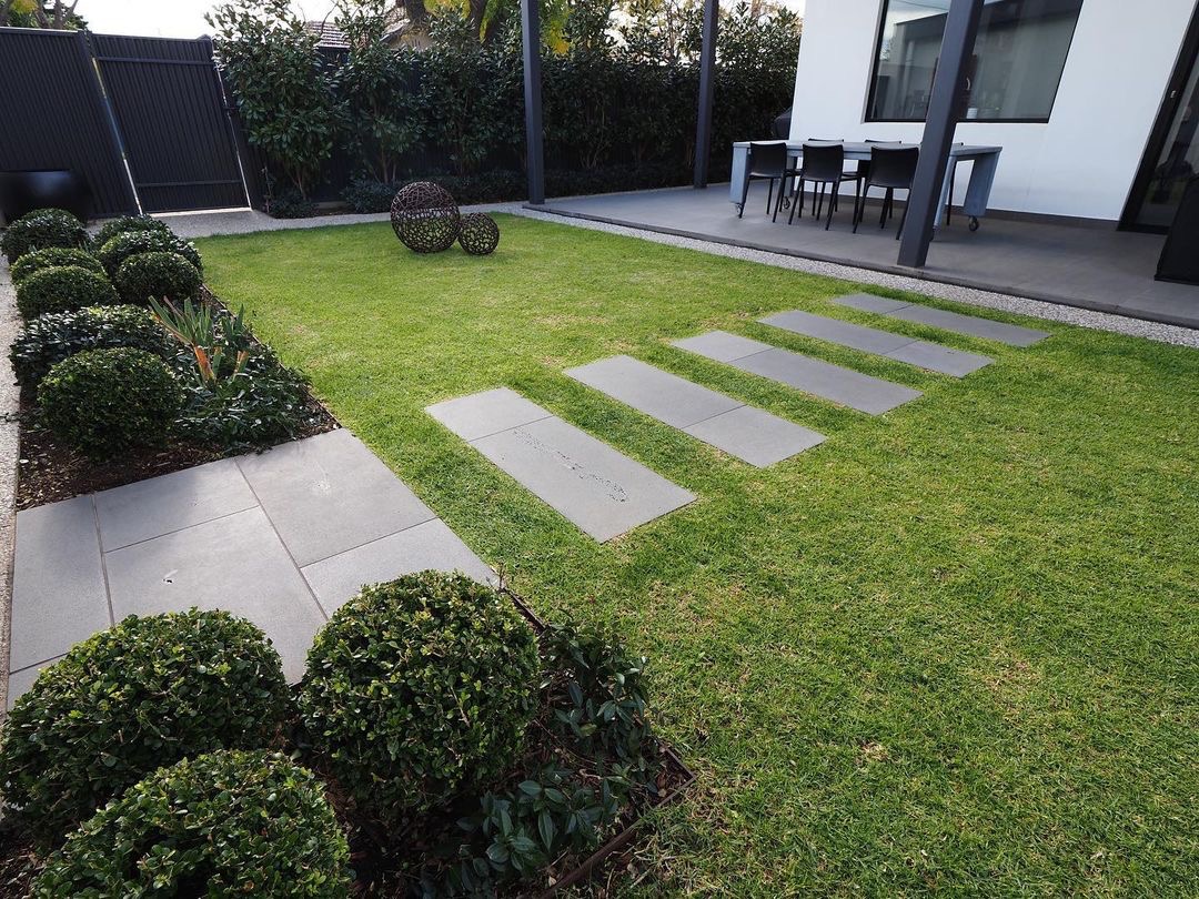 Nowoczesny ogród z równo przystrzyżonym trawnikiem, ścieżką z płyt betonowych, kulistymi krzewami bukszpanu i dekoracyjnymi kulami z metalu.