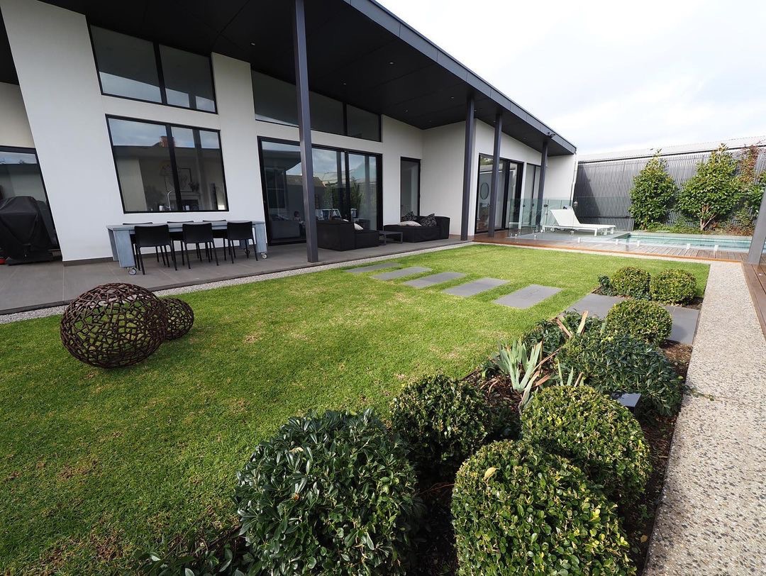 Nowoczesny ogród z równo przystrzyżonym trawnikiem, ścieżką z płyt kamiennych, dekoracyjnymi kulami z wikliny i basenem w tle, otoczony niskimi, kulistymi krzewami.