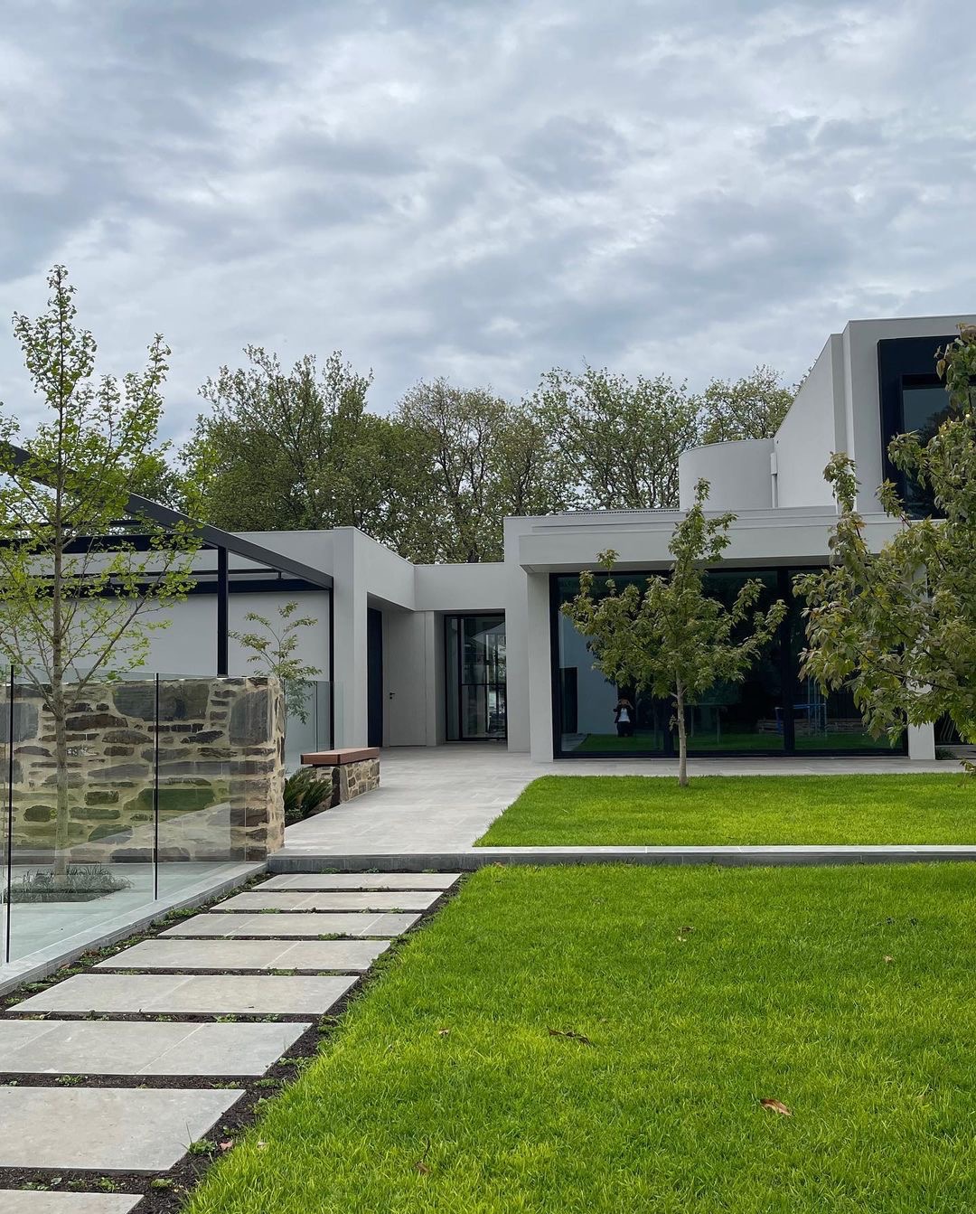 Nowoczesny dom z zadbanym trawnikiem, ścieżką z płyt betonowych i elementami małej architektury ogrodowej, takimi jak kamienny murek i przeszklona pergola.