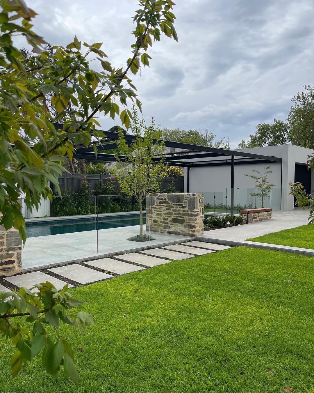 Nowoczesny ogród z równo przystrzyżonym trawnikiem, basenem otoczonym szklanym ogrodzeniem, kamiennymi słupkami i ścieżką z płyt. W tle pergola i budynek w minimalistycznym stylu.