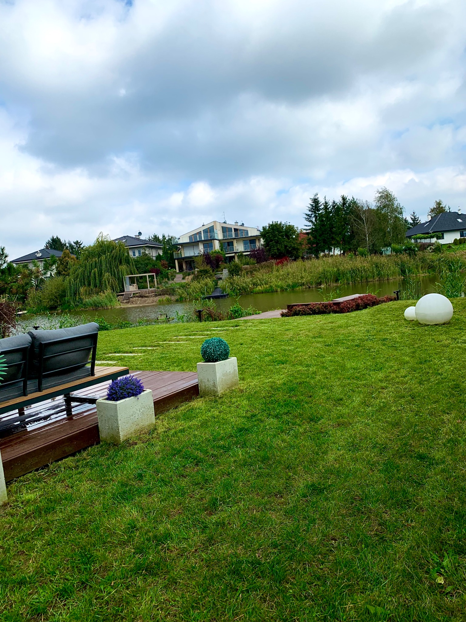 Widok na zadbany trawnik z dekoracjami ogrodowymi, w tle jezioro i nowoczesne domy pod pochmurnym niebem.