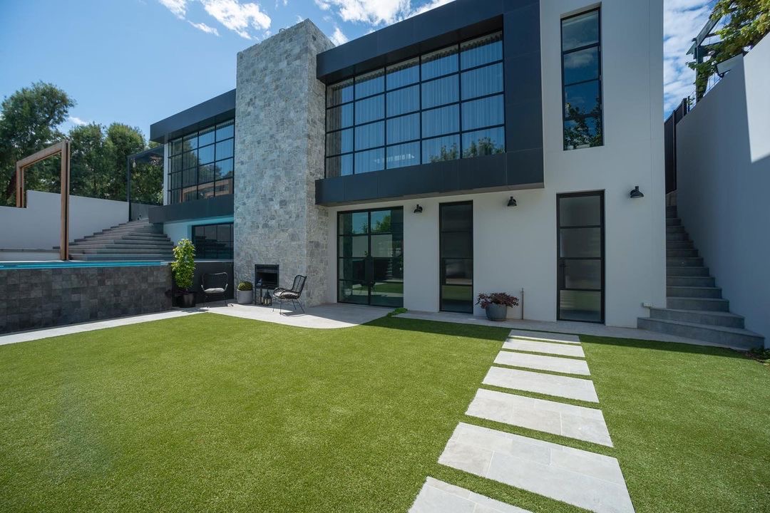 Nowoczesny dom z dużymi oknami i sztuczną trawą, z wyłożonymi płytami chodnikowymi prowadzącymi do wejścia, obok basenu i schodów.