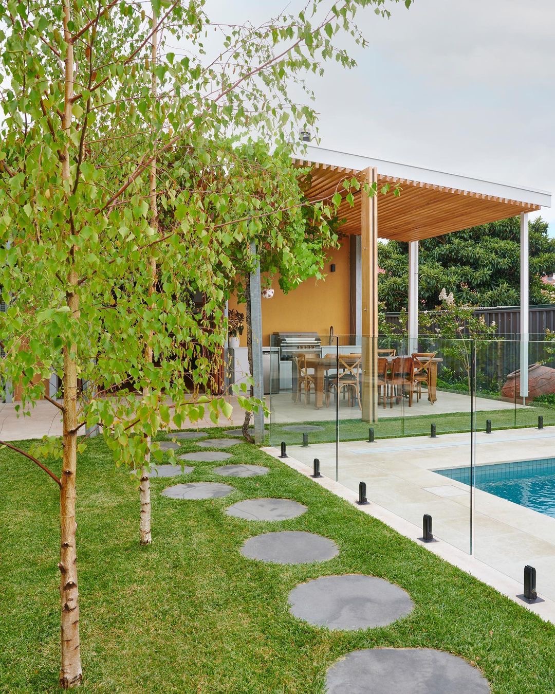 Nowoczesny ogród z trawnikiem, ścieżką z okrągłych płyt, młodymi brzozami i basenem otoczonym szklanym ogrodzeniem, zadaszona altana z drewnianymi elementami w tle.