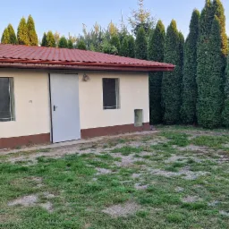 Niewielki, otynkowany budynek z czerwoną dachówką i brązową podmurówką, otoczony tujami, na trawiastym terenie.