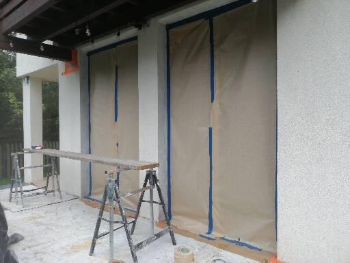 Elewacja budynku zabezpieczona folią i niebieską taśmą malarską przed pracami, widoczne rusztowania i częściowo pomalowana ściana.