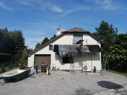 Dom jednorodzinny z rusztowaniem podczas prac elewacyjnych; widoczna plandeka ochronna, drabina i sprzęt budowlany na podjeździe z kostki brukowej.