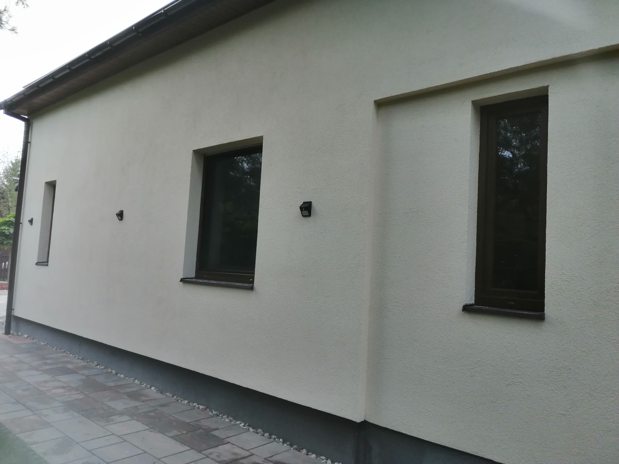 Jasna elewacja domu jednorodzinnego z ciemnymi oknami i oświetleniem zewnętrznym, widok z boku na fragment ściany i chodnik z kostki brukowej.