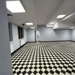 Wnętrze biura po remoncie: szara ściana, czarno-biała podłoga w szachownicę, białe grzejniki i sufit z kwadratowymi lampami LED.