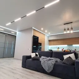 Nowoczesne wnętrze salonu z aneksem kuchennym, oświetlenie listwowe LED na suficie, szara sofa z poduszkami i pledem, drewniane blaty kuchenne, przesuwne drzwi do szafy w tle.