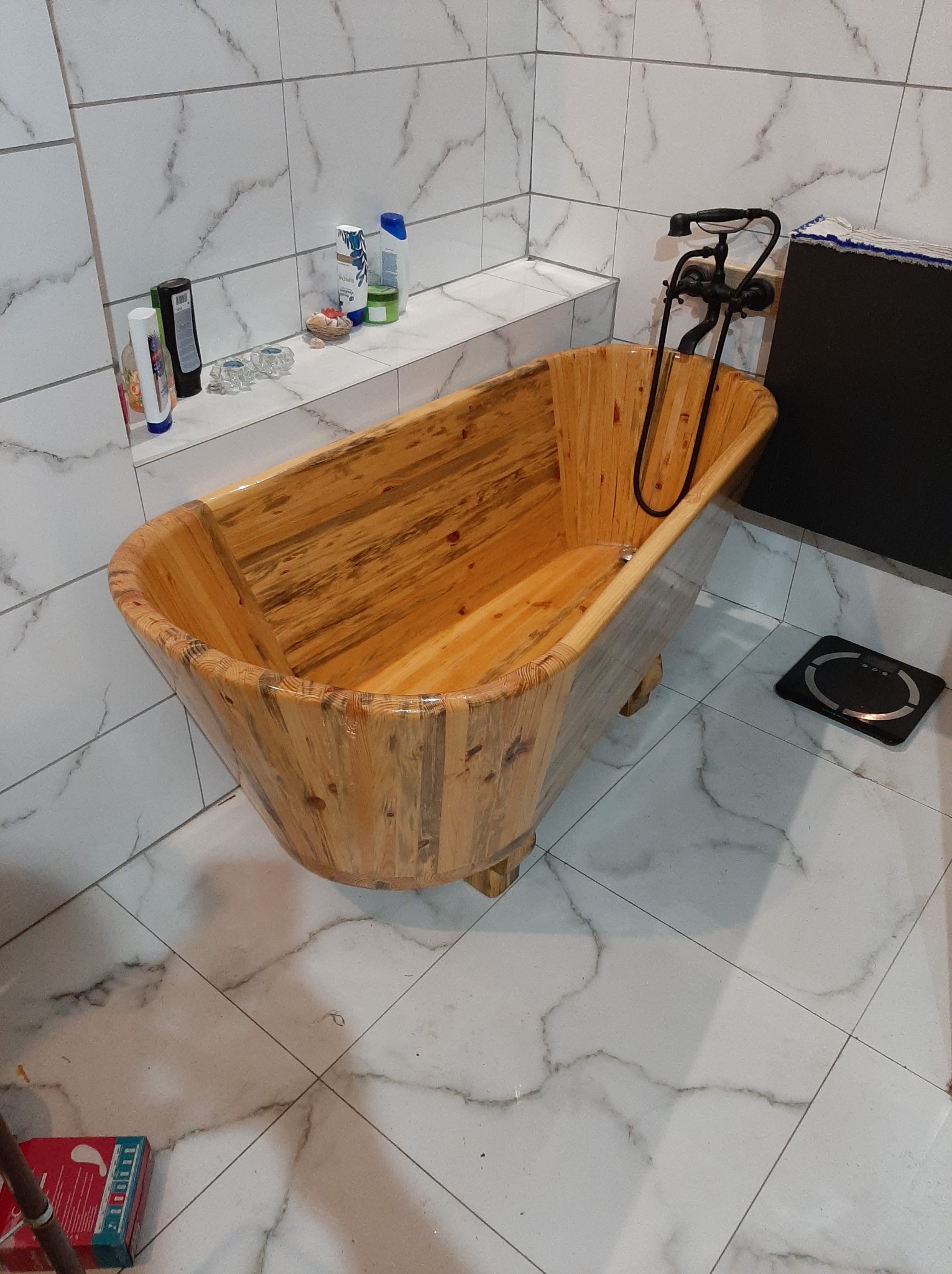 Rustykalna wanna z jasnego drewna z czarną armaturą na tle białych płytek imitujących marmur. Widoczna półka z kosmetykami i waga łazienkowa.