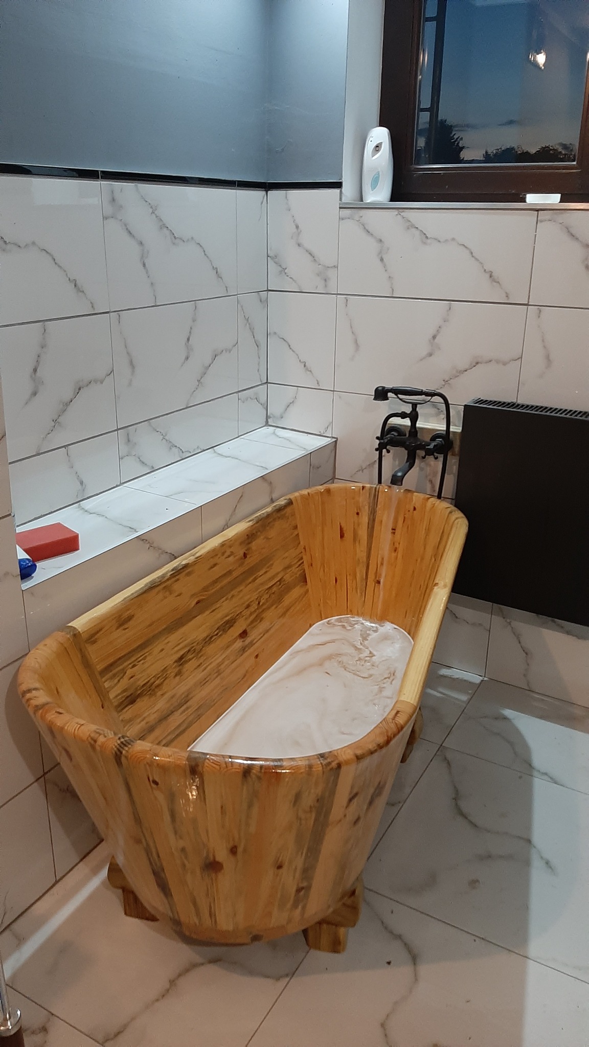 Drewniana wanna z czarną armaturą w łazience wyłożonej białymi płytkami z marmurkowym wzorem.