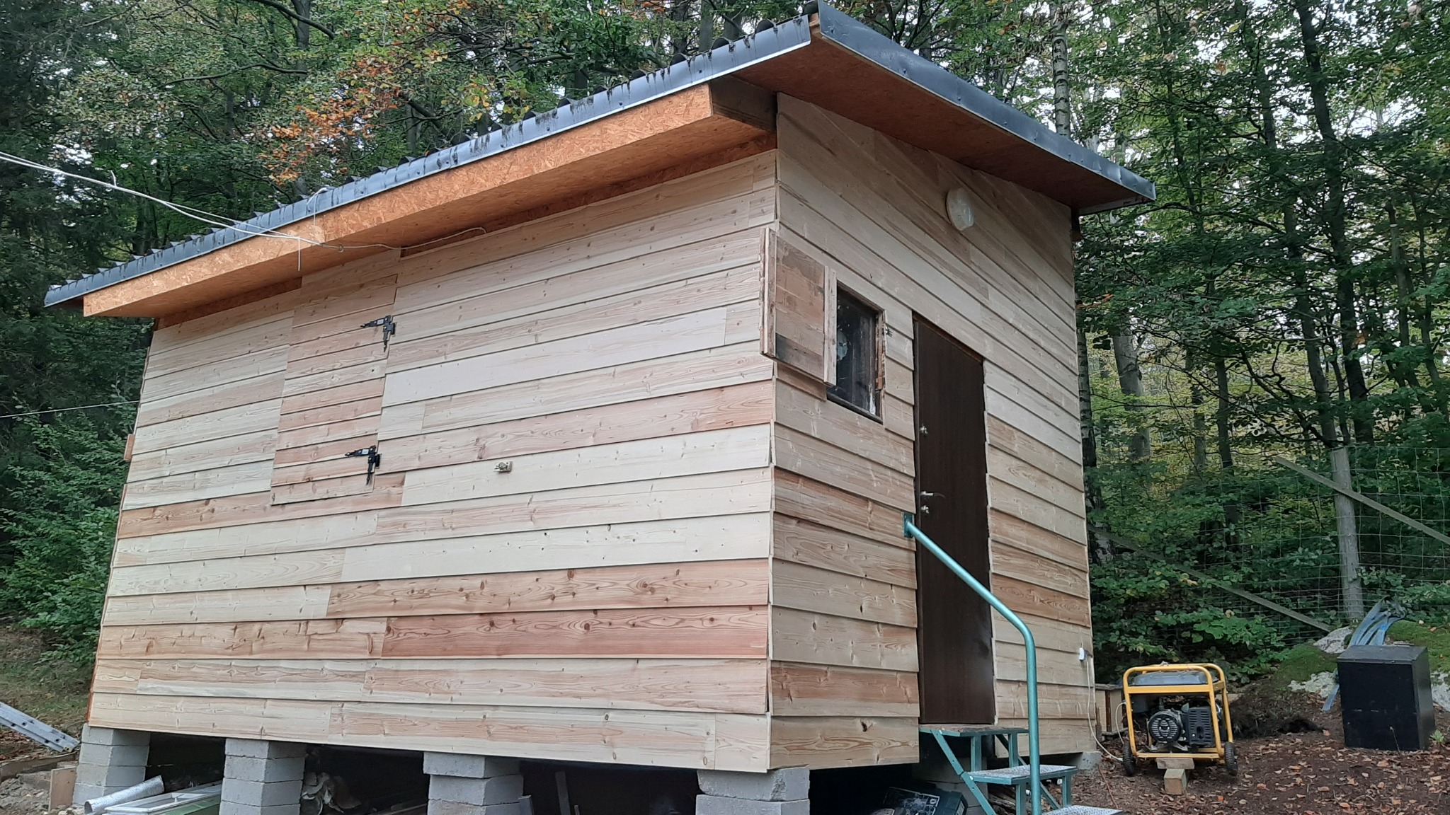 Drewniany domek gospodarczy z ciemnymi drzwiami, oknem z okiennicą, na betonowych bloczkach, w tle las i generator prądu.