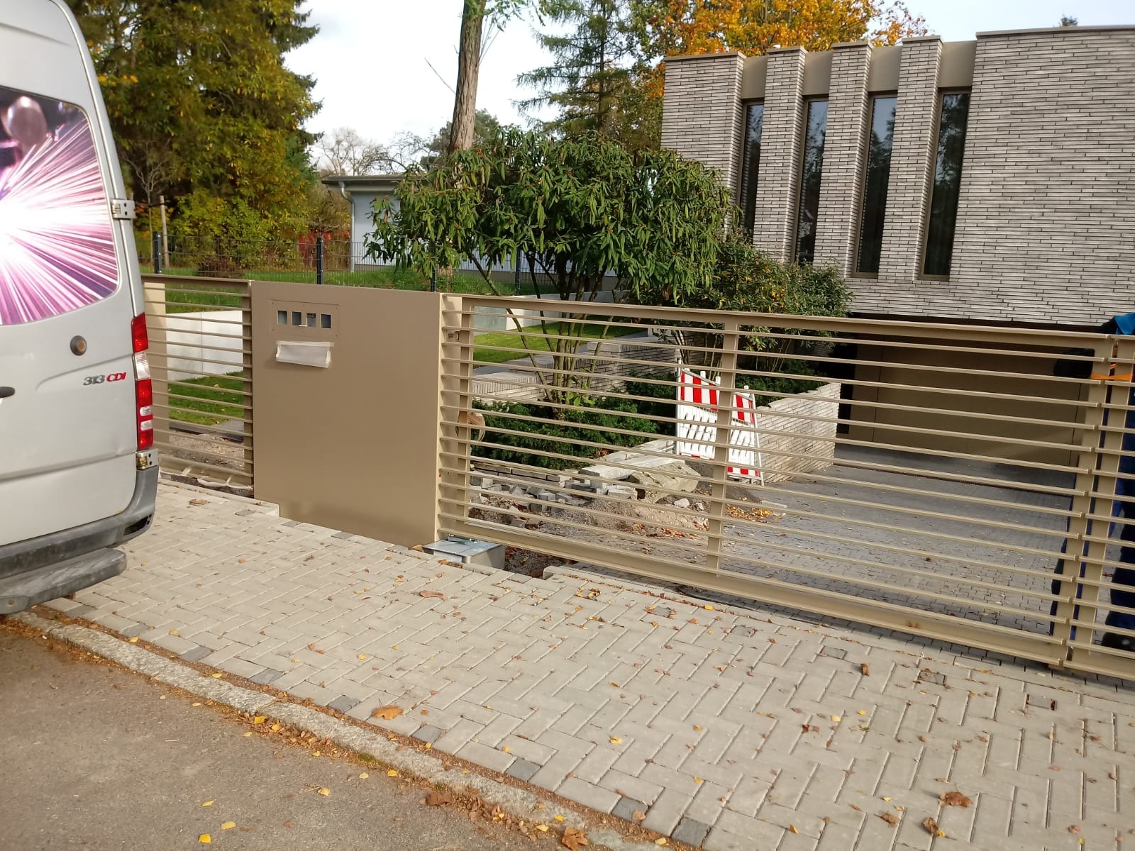 Automatyczna brama posesyjna z poziomych profili, zintegrowana ze skrzynką pocztową, w trakcie montażu, widoczne zabezpieczenia budowlane i samochód dostawczy.