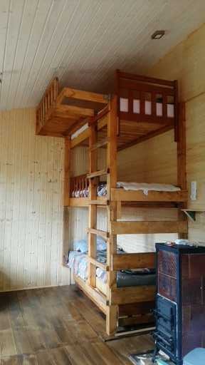 Trzypoziomowe drewniane łóżko piętrowe z drabinką, w pokoju wykończonym boazerią, z piecem kaflowym w rogu.