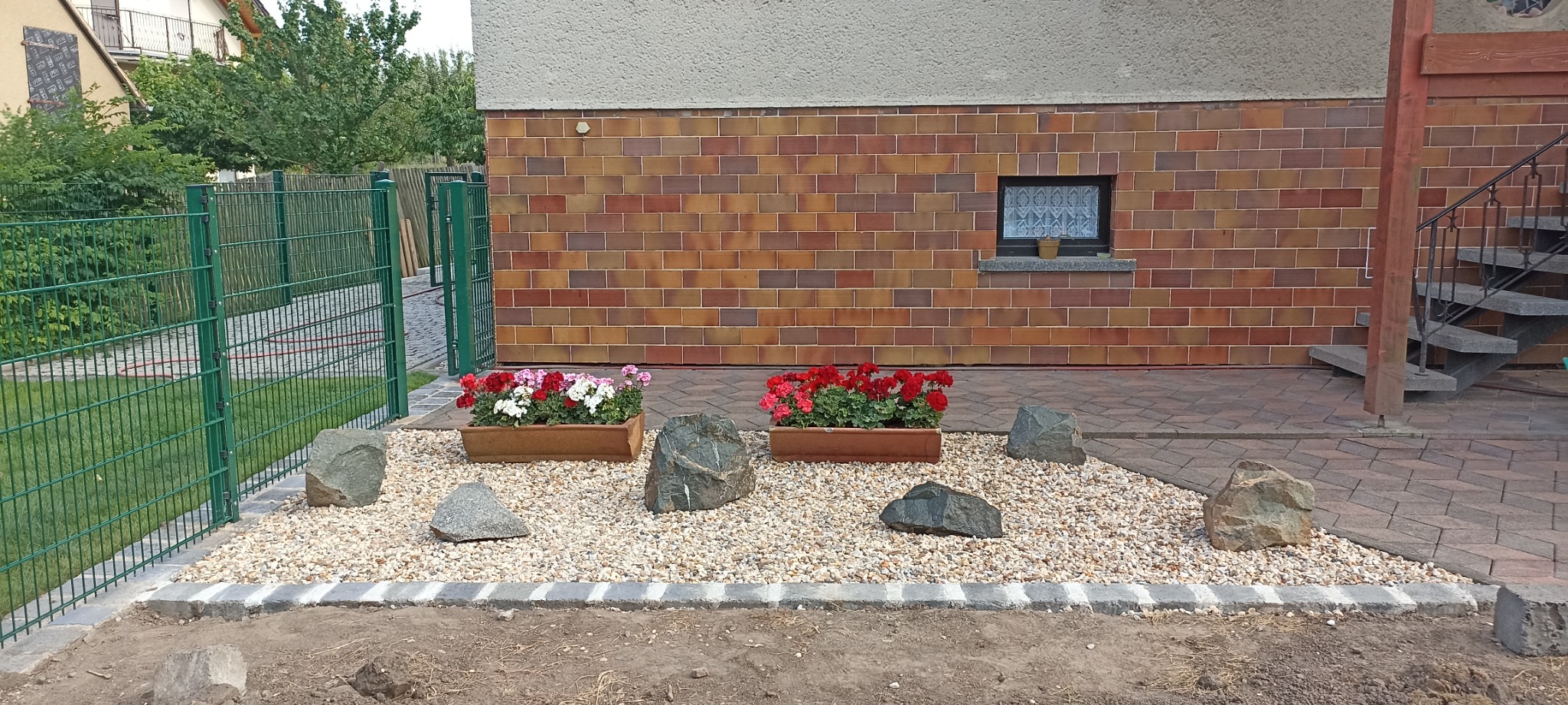 Ogród przed domem z zielonym ogrodzeniem panelowym, rabatą z kamyków, donicami z kwiatami i dużymi kamieniami dekoracyjnymi.