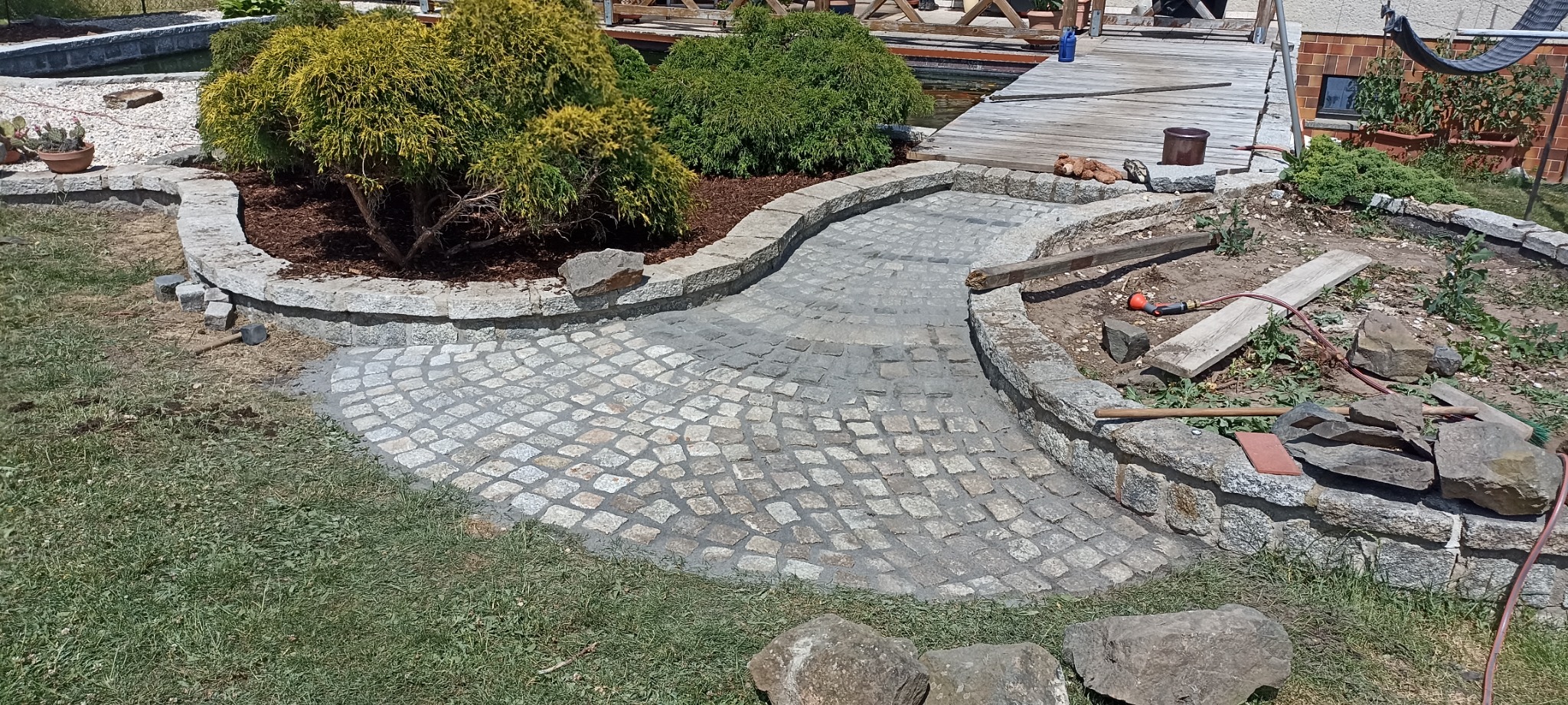 Wykonanie ścieżki z kostki granitowej w ogrodzie, otoczonej kamiennym murkiem, z widocznymi narzędziami i elementami krajobrazu.