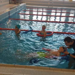 Instruktor w czarnym stroju pomaga dziecku utrzymać się na wodzie na basenie; w tle inne dzieci z makaronami do pływania.