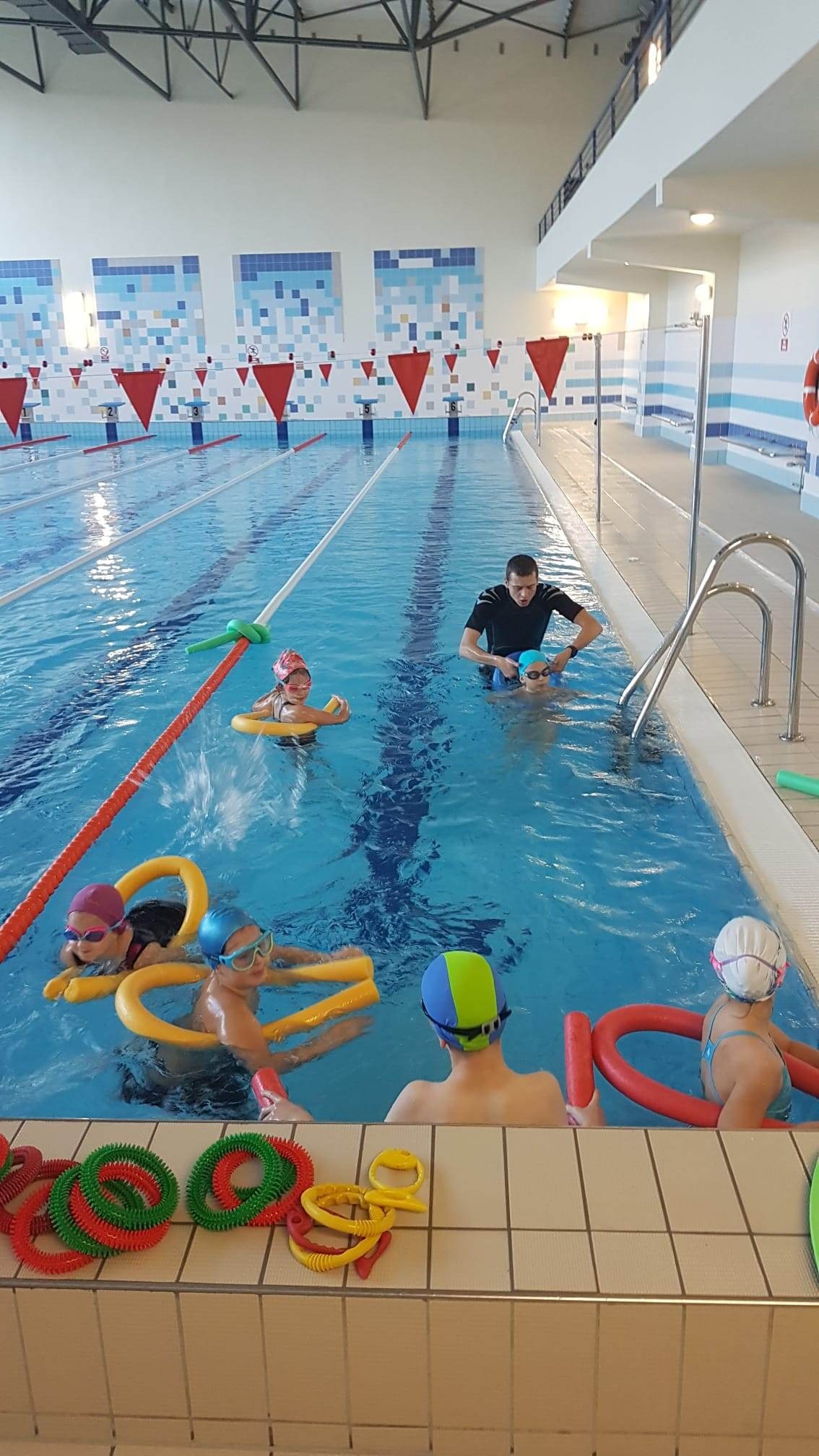 Instruktor w czarnej koszulce pomaga dziecku w niebieskim czepku na basenie, w tle dzieci z makaronami pływackimi i kołami, kolorowe zabawki na brzegu basenu.