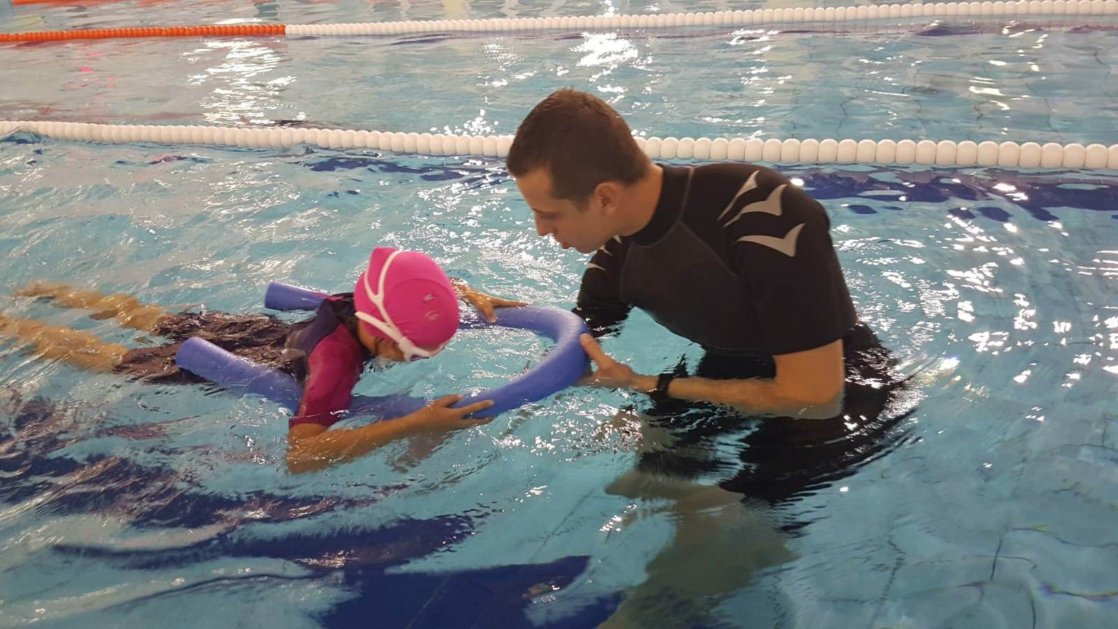 Instruktor w czarnej koszulce z długim rękawem i wzorem na ramieniu pomaga dziecku w różowym czepku pływać z piankową rurą w basenie.