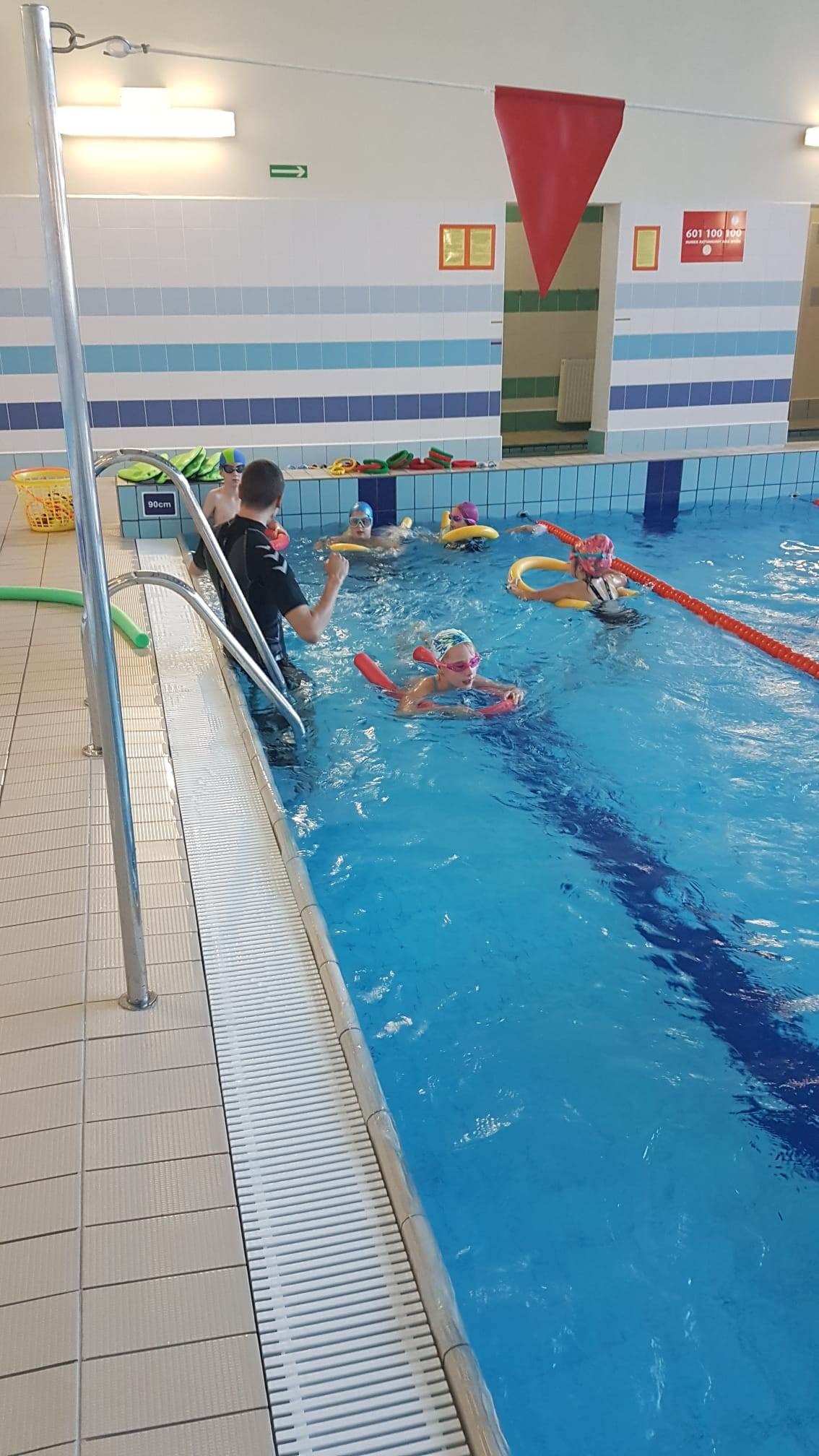 Instruktor prowadzi zajęcia w basenie z grupą dzieci, które uczą się pływać z użyciem piankowych makaronów i kółek, widoczna płytka woda i oznaczenia głębokości.