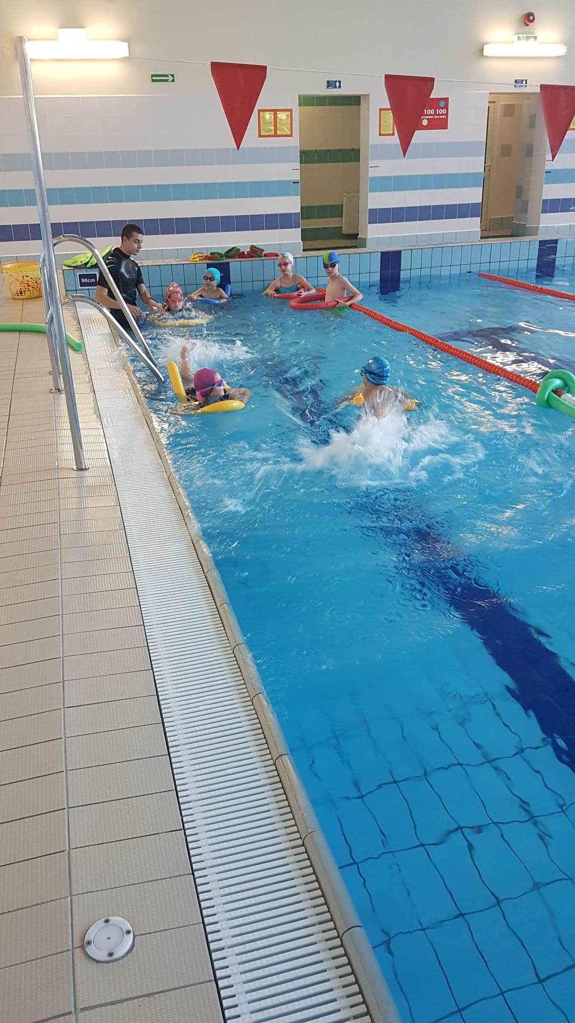 Instruktor prowadzi zajęcia z pływania dla grupy dzieci na basenie we Wrocławiu. Dzieci w czepkach i z akcesoriami do pływania, w tle czerwone flagi.
