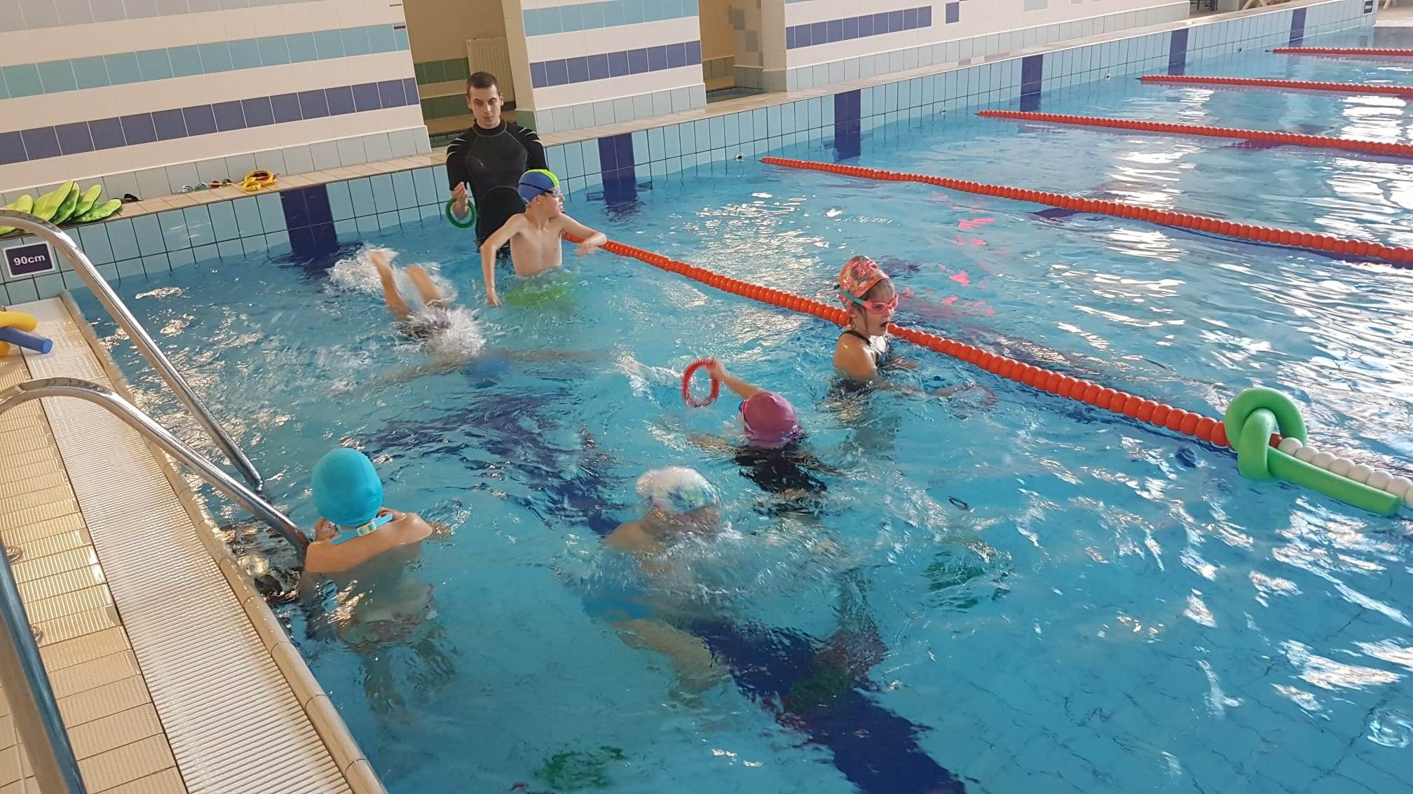 Grupowe zajęcia na basenie: instruktor w czarnym stroju nadzoruje dzieci w czepkach i okularach pływackich, używających piankowych makaronów i kółek do nauki pływania w płytkim basenie...