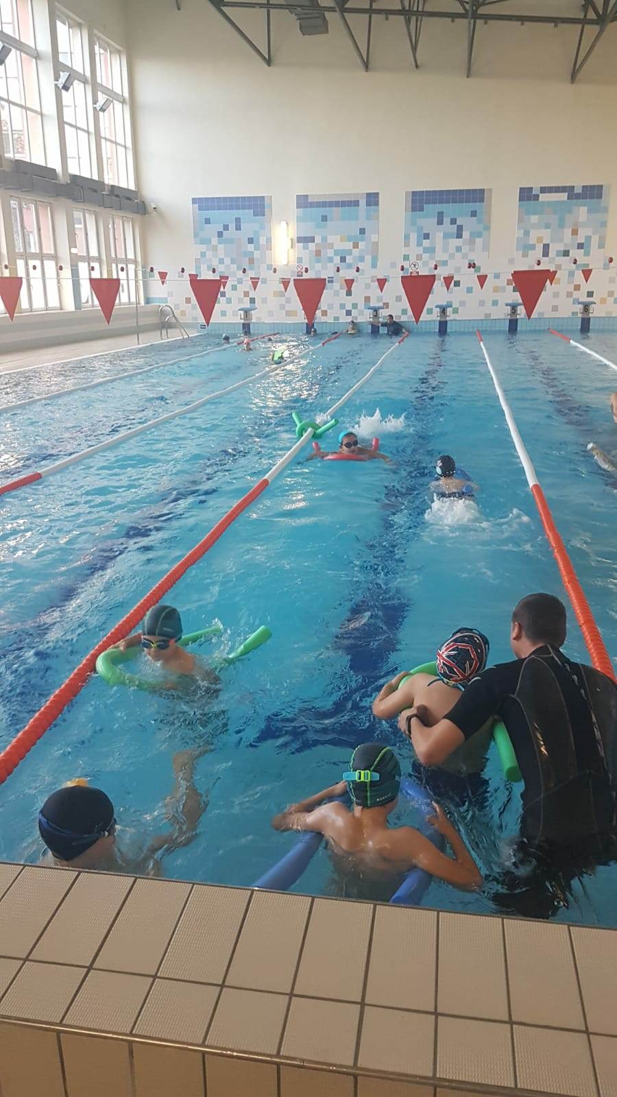 Grupa dzieci w czepkach i okularach pływackich ćwiczy pływanie na basenie pod okiem instruktora, używając piankowych makaronów.