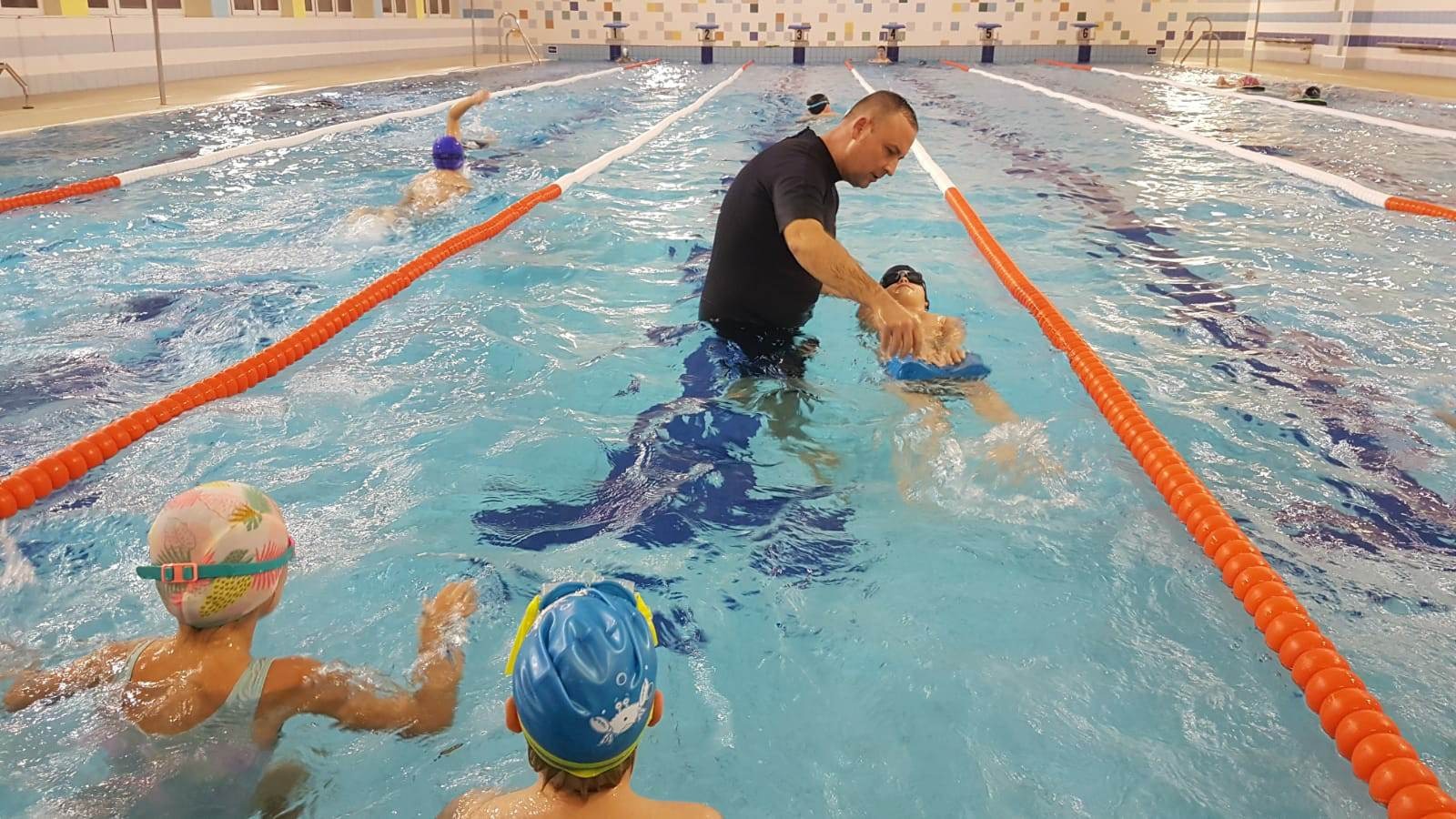 Instruktor w czarnym stroju asekuruje dziecko z niebieską deską podczas lekcji pływania na basenie, w tle widoczni inni uczestnicy zajęć.