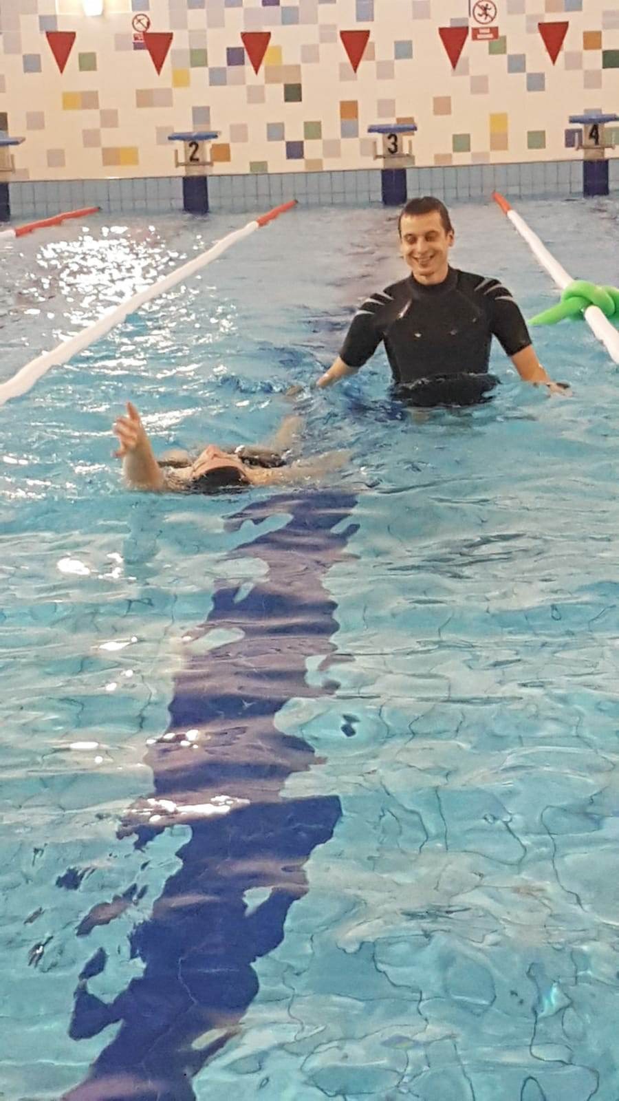 Instruktor w czarnej piance uśmiecha się, stojąc w basenie obok kobiety unoszącej rękę w górę podczas lekcji pływania na tle kafelkowej ściany z oznaczeniami torów.