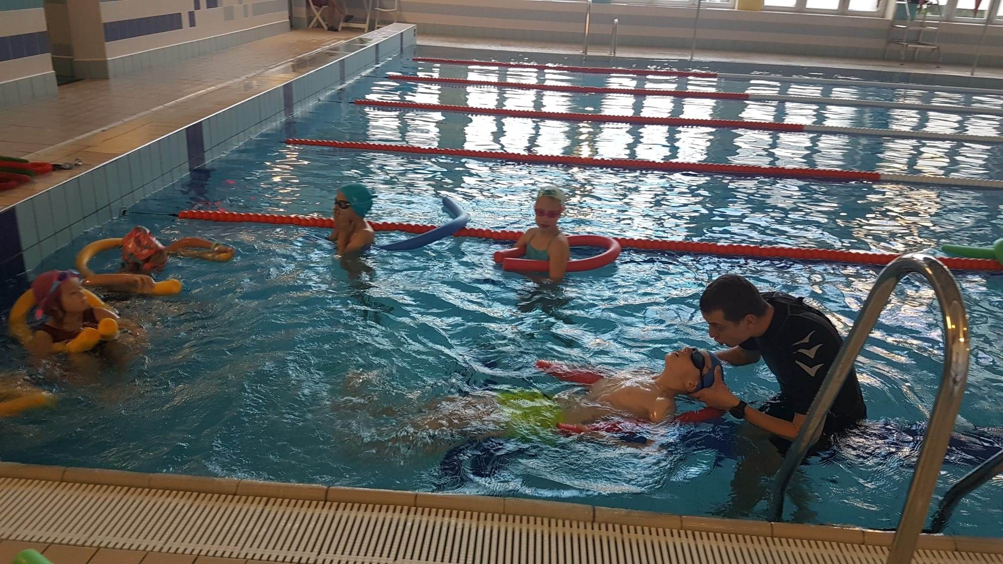 Instruktor w czarnym stroju pomaga dziecku utrzymać się na wodzie na basenie; w tle inne dzieci z makaronami do pływania.