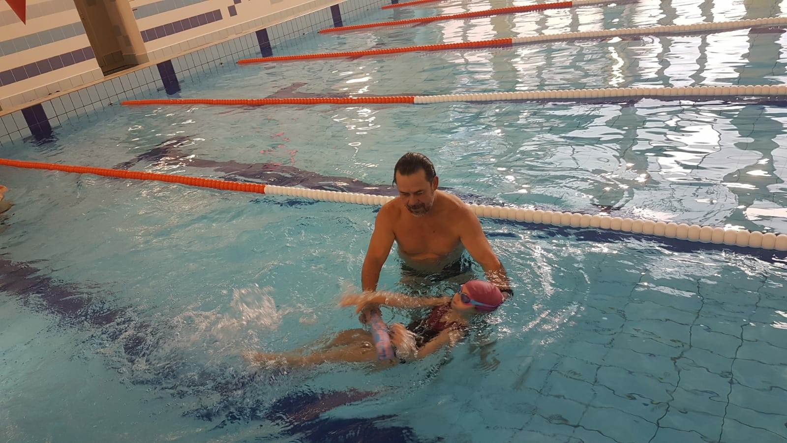 Instruktor pomaga dziewczynce w różowym czepku unosić się na wodzie w basenie z niebieskimi i białymi pasami na ścianach.