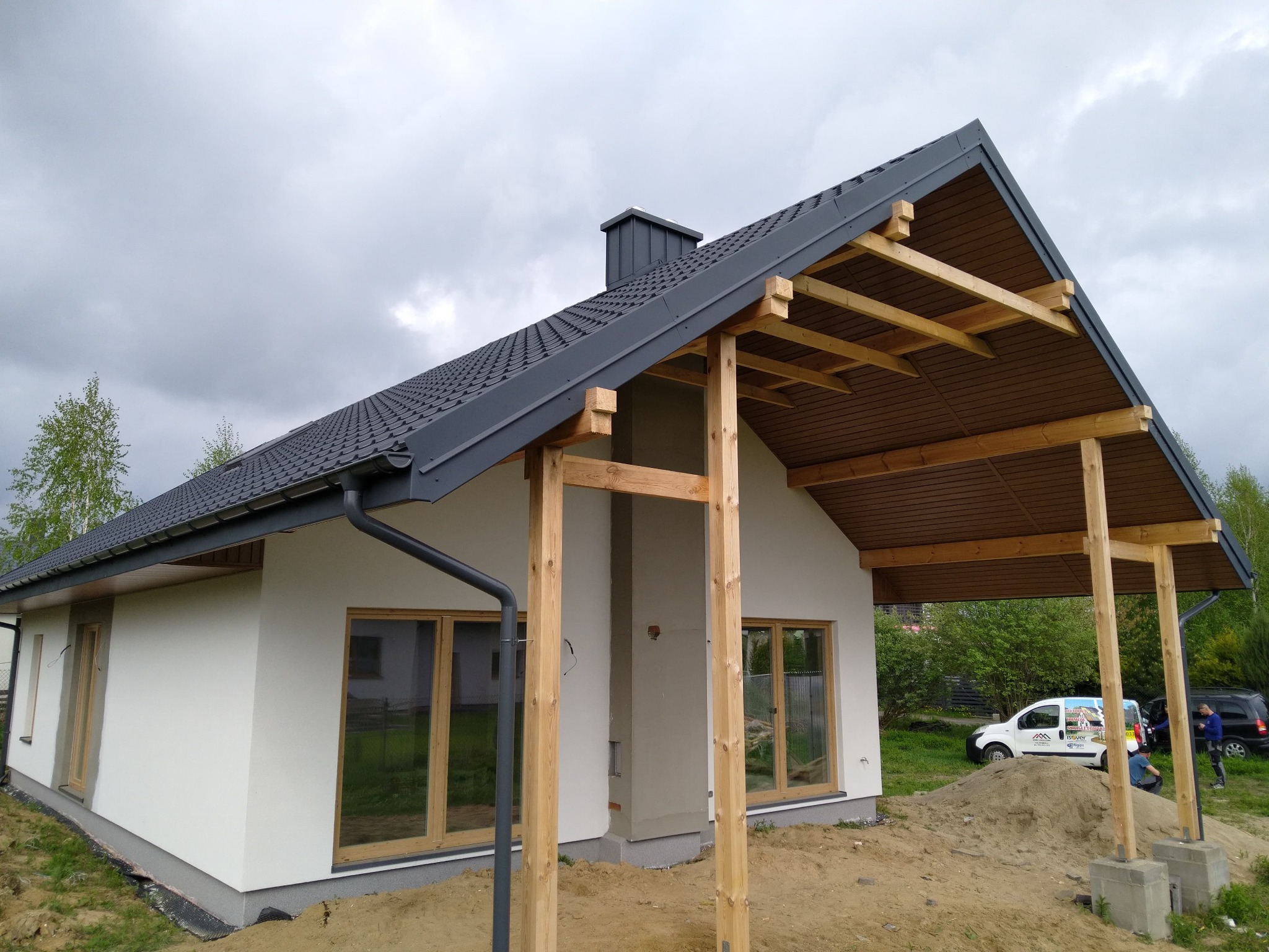 Nowoczesny dom jednorodzinny z szarą dachówką, drewnianą konstrukcją zadaszenia tarasu i świeżo otynkowaną, białą elewacją w trakcie prac wykończeniowych, z widocznym stosem piasku i samochodem...